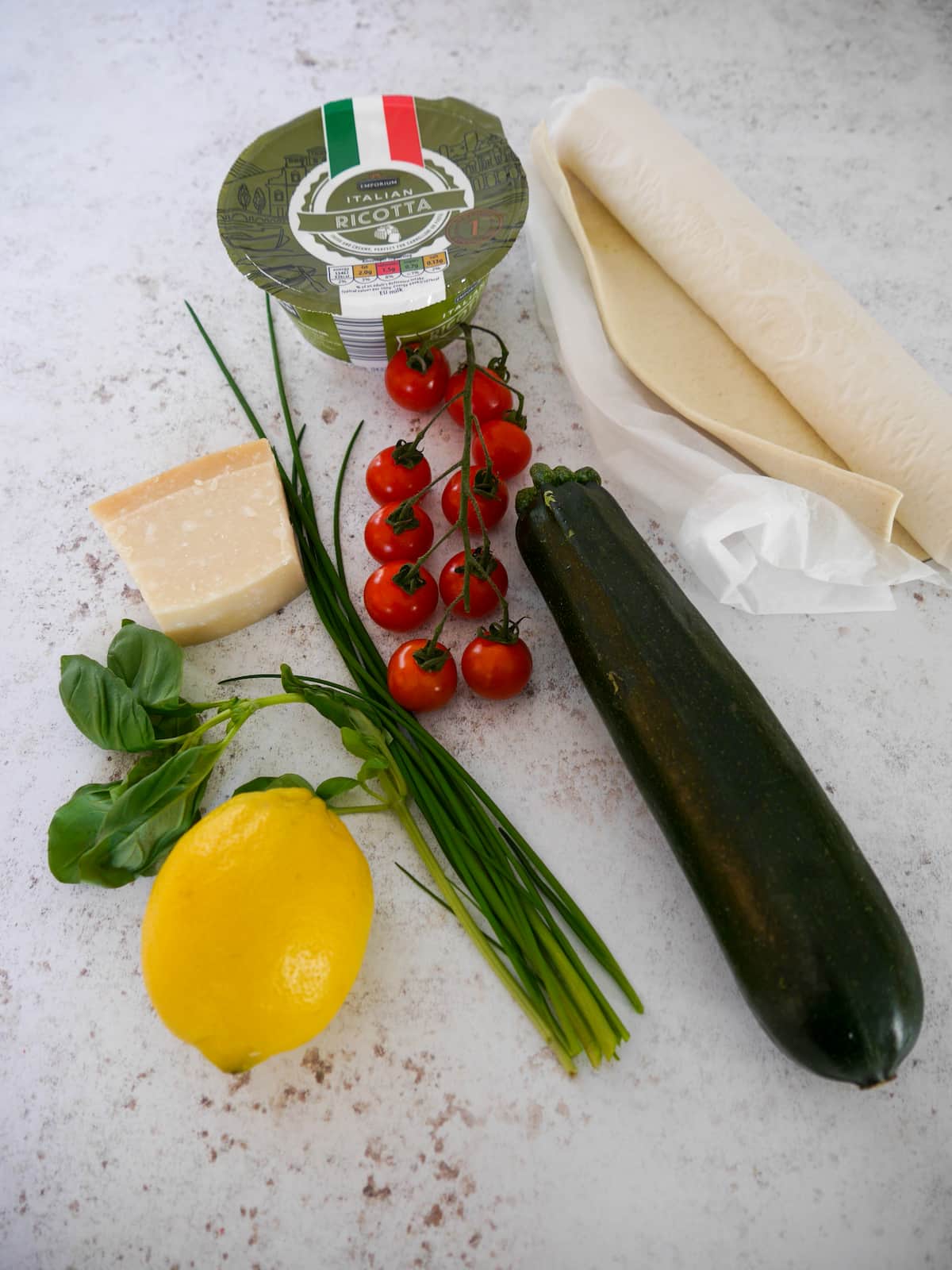 Individual recipe ingredients for making tomato and courgette puff pastry tart with ricotta cheese, fresh herbs and lemon.