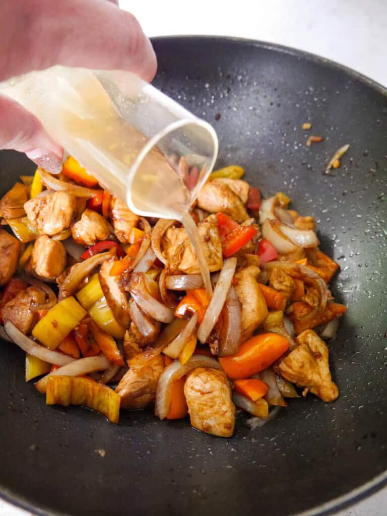 Chicken stock being poured over a wok of stir-fried chicken and vegetables.