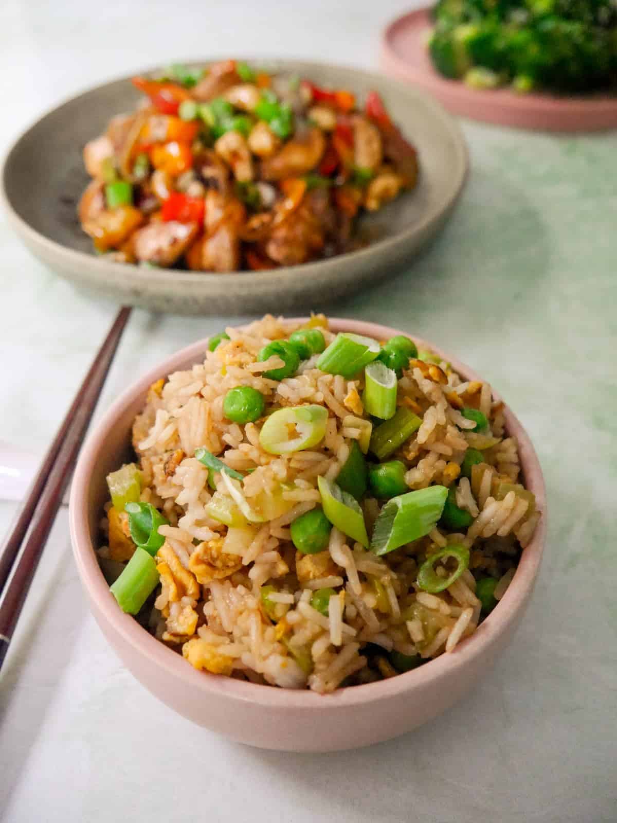 A bowl of egg fried rice, garnished with sliced spring onions, with bowls of stir-fried chicken, and stir-fried broccoli set alongside.