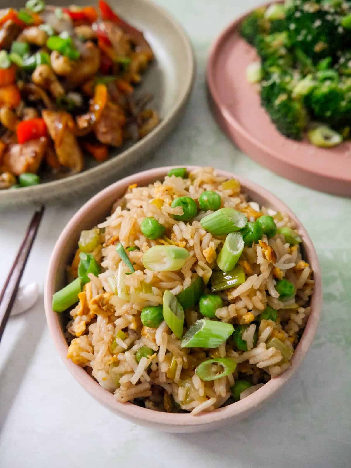 A bowl of egg fried rice, garnished with sliced spring onions, with bowls of stir-fried chicken, and stir-fried broccoli set alongside.