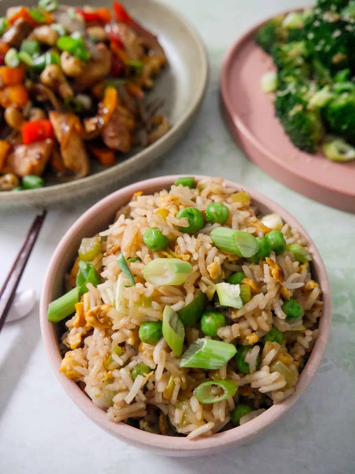 A bowl of egg fried rice, garnished with sliced spring onions, with bowls of stir-fried chicken, and stir-fried broccoli set alongside.