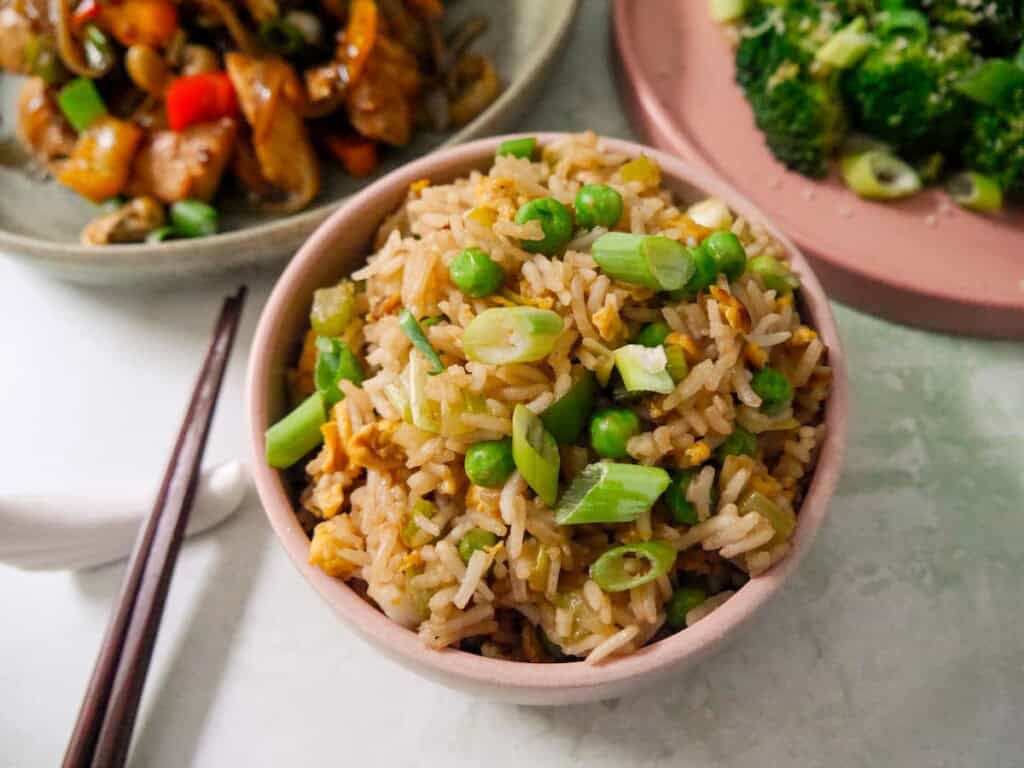 A bowl of egg fried rice, garnished with sliced spring onions, with bowls of stir-fried chicken, and stir-fried broccoli set alongside.