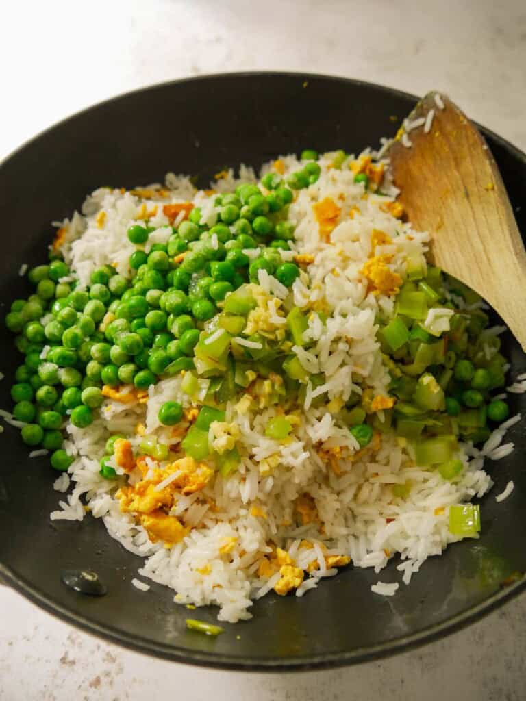 A wok of egg fried rice with sauteed leek and celery being added back into the wok and frozen garden peas being added.