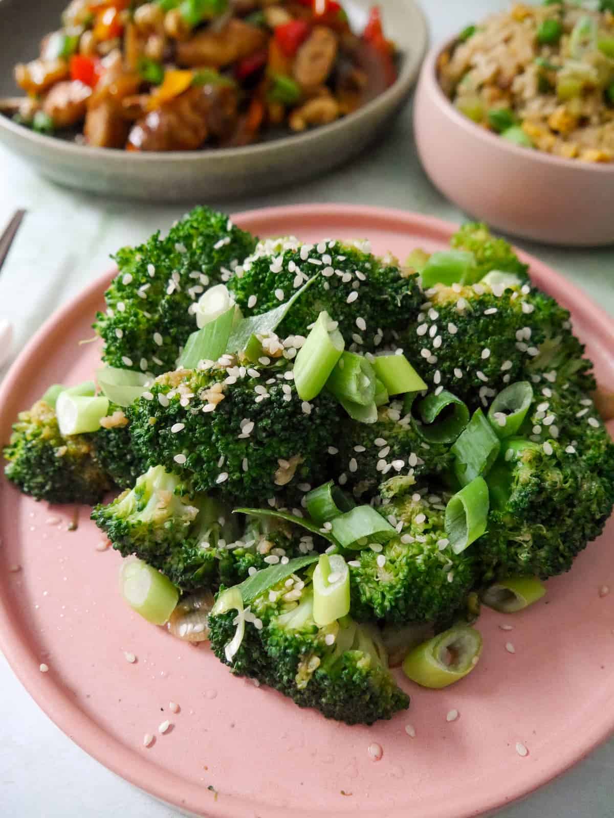A plate of stir-fried broccoli garnished with sesame seeds and sliced spring onions, with bowls of stir-fried chicken and egg fried rice set alongside.
