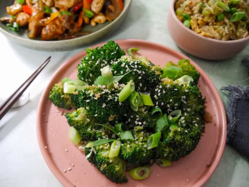 A plate of stir-fried broccoli garnished with sesame seeds and sliced spring onions, with bowls of stir-fried chicken and egg fried rice set alongside.
