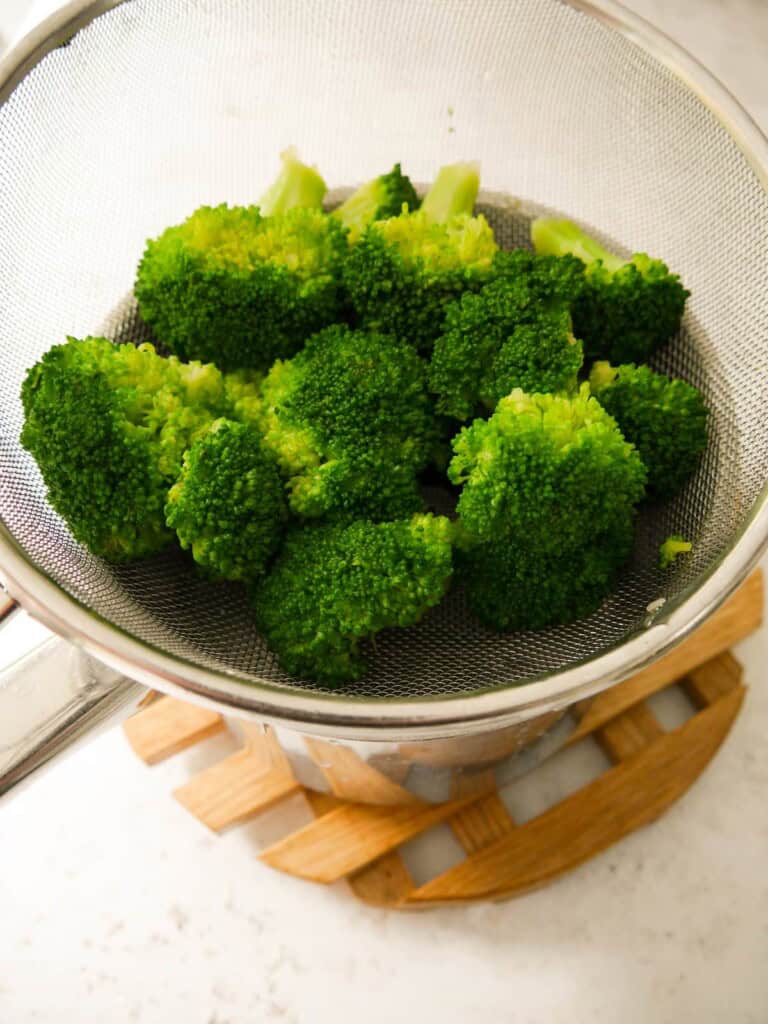 Blanched broccoli florets set in a sieve over a saucepan to steam dry.