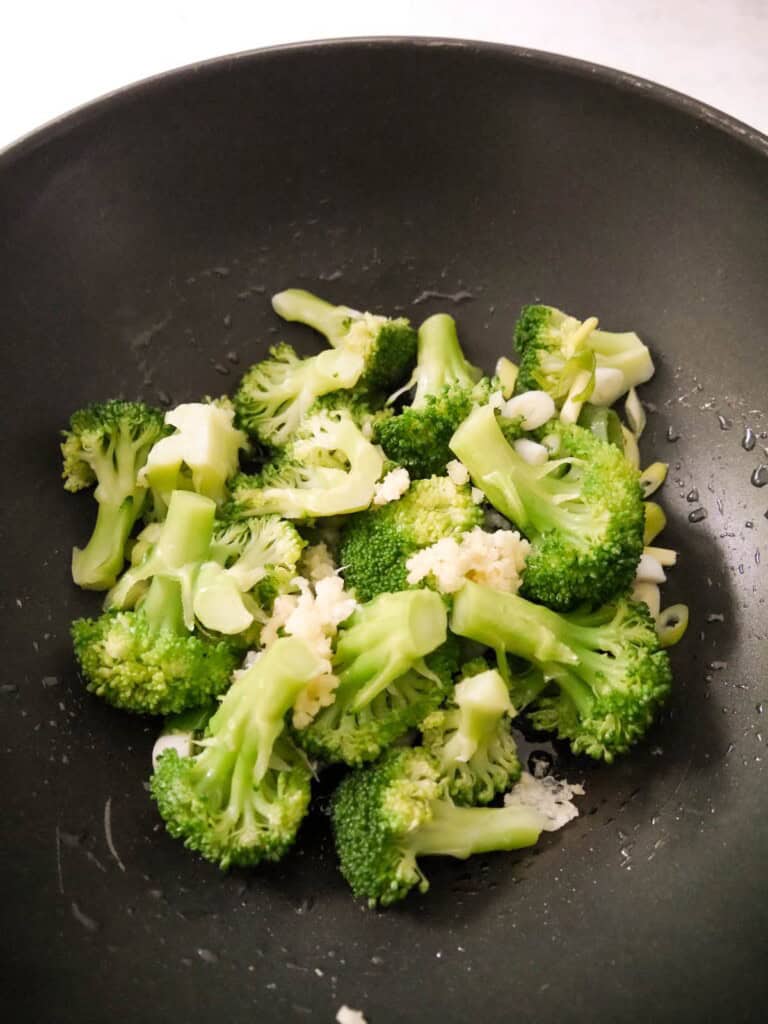 A wok with blanched broccoli florets with added crushed garlic and sliced spring onions.