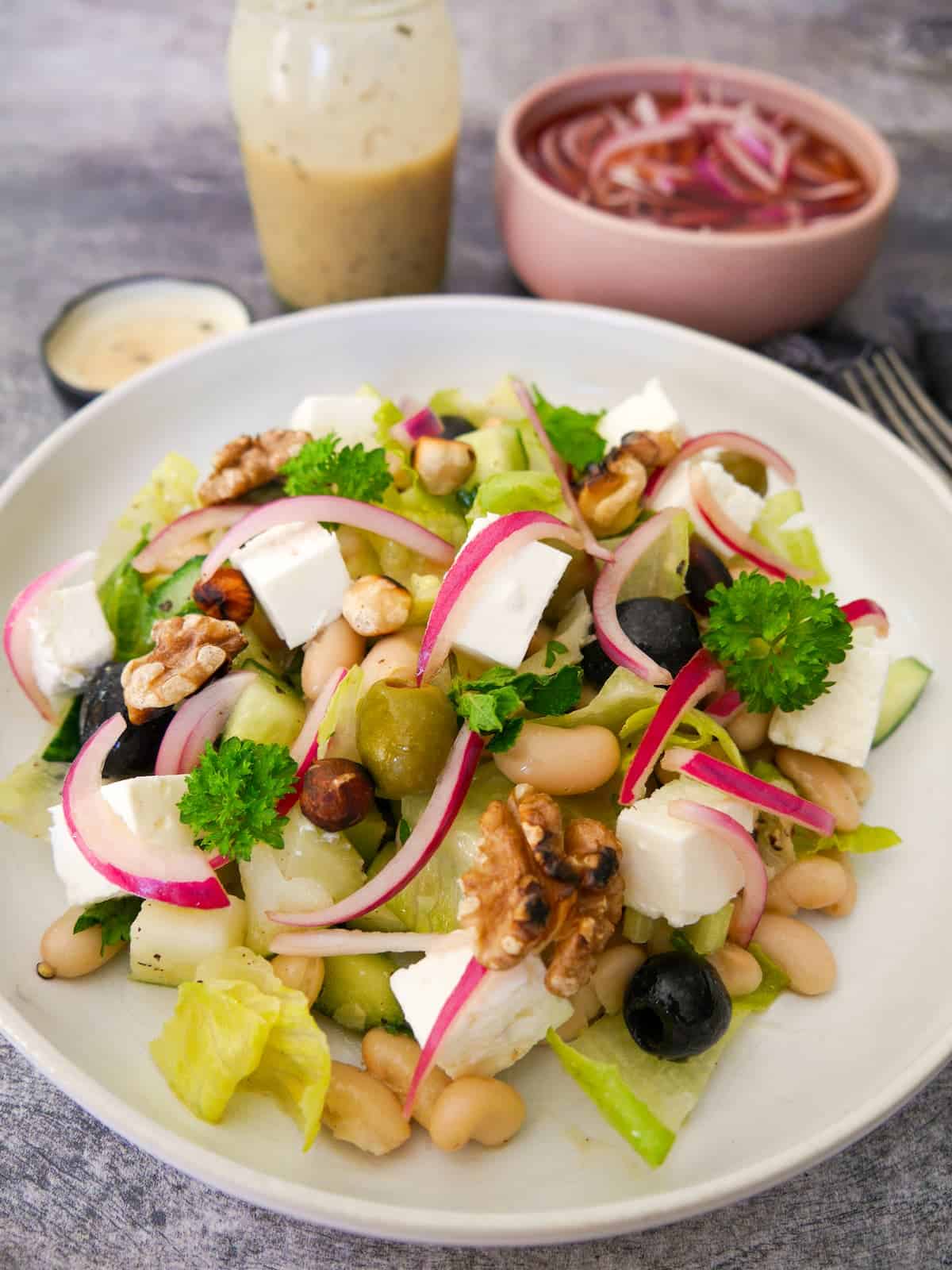 A bowl of white bean and feta salad, which olives, pickled red onions and toasted nuts, with a bowl of red onions and salad dressing set alongside.