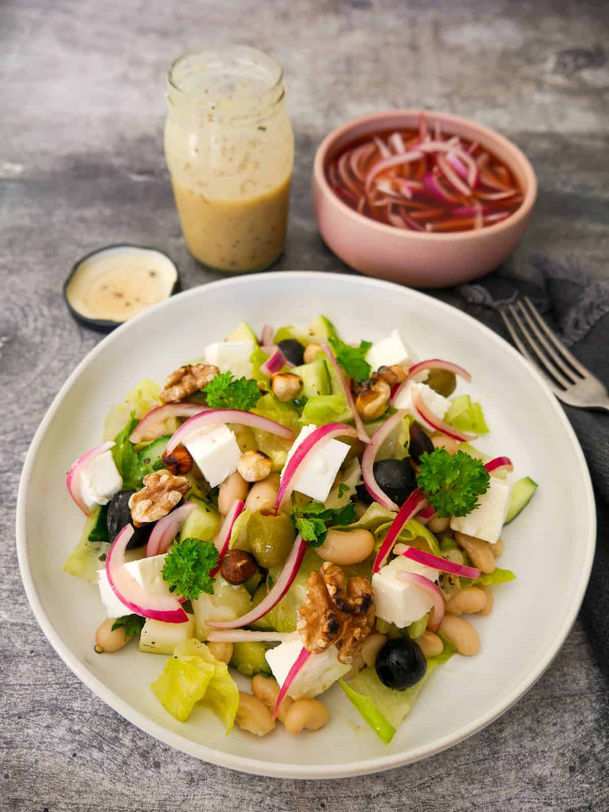 A bowl of white bean and feta salad, which olives, pickled red onions and toasted nuts, with a bowl of red onions and salad dressing set alongside.