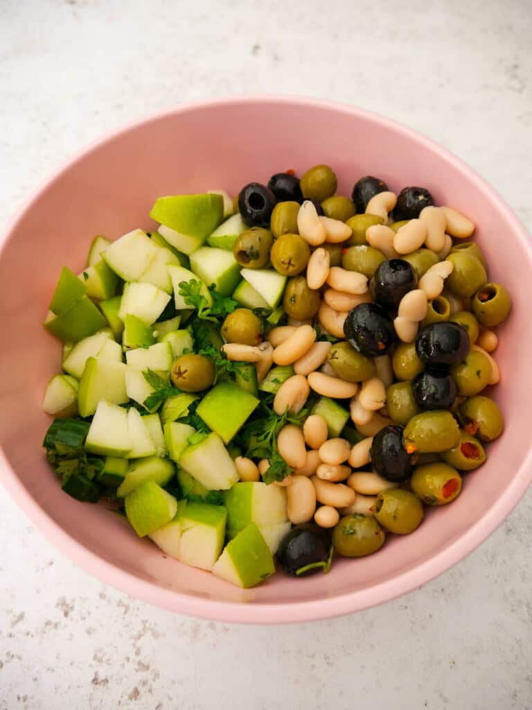 A bowl of mixed salad ingredients including white beans, mixed olives, green apple, celery and freshly chopped herbs.