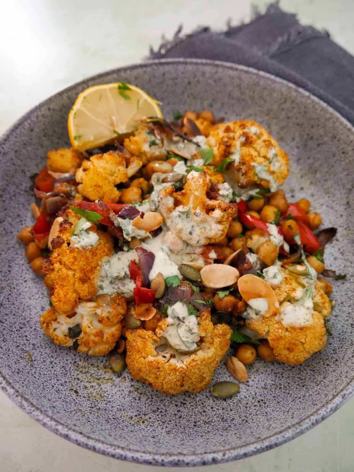 A bowl of roasted cauliflower and chickpeas, drizzled with Greek yogurt lemon herb dressing, and garnished with fresh herbs, and toasted almonds and pumpkin seeds.