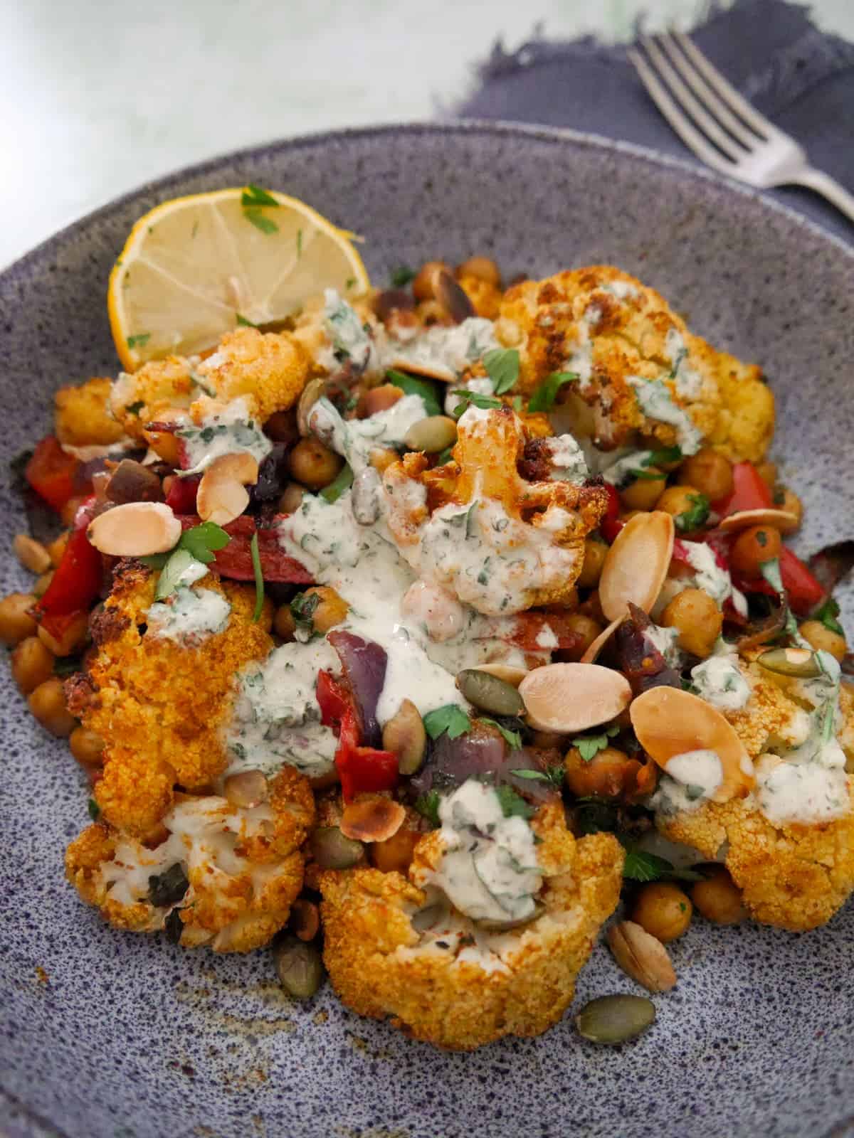 A bowl of roasted cauliflower and chickpeas, drizzled with Greek yogurt lemon herb dressing, and garnished with fresh herbs, and toasted almonds and pumpkin seeds.