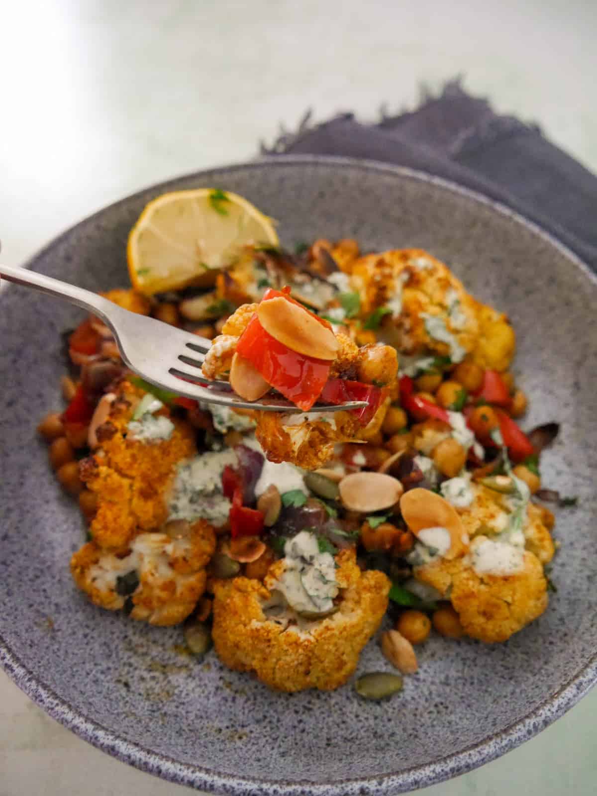 A bowl of roasted cauliflower and chickpeas, drizzled with Greek yogurt lemon herb dressing, and garnished with fresh herbs, and toasted almonds and pumpkin seeds.