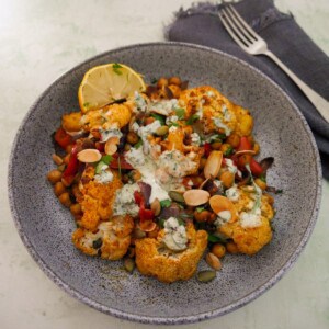 A bowl of roasted cauliflower and chickpeas, drizzled with Greek yogurt lemon herb dressing, and garnished with fresh herbs, and toasted almonds and pumpkin seeds.