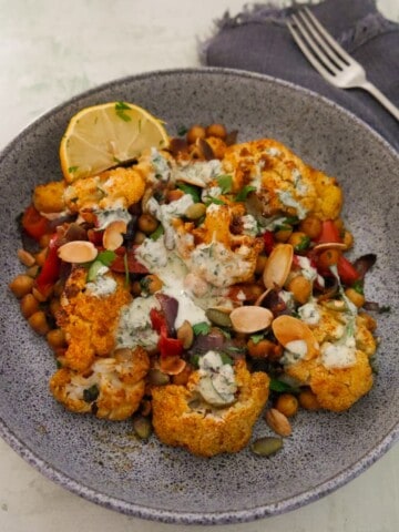 A bowl of roasted cauliflower and chickpeas, drizzled with Greek yogurt lemon herb dressing, and garnished with fresh herbs, and toasted almonds and pumpkin seeds.