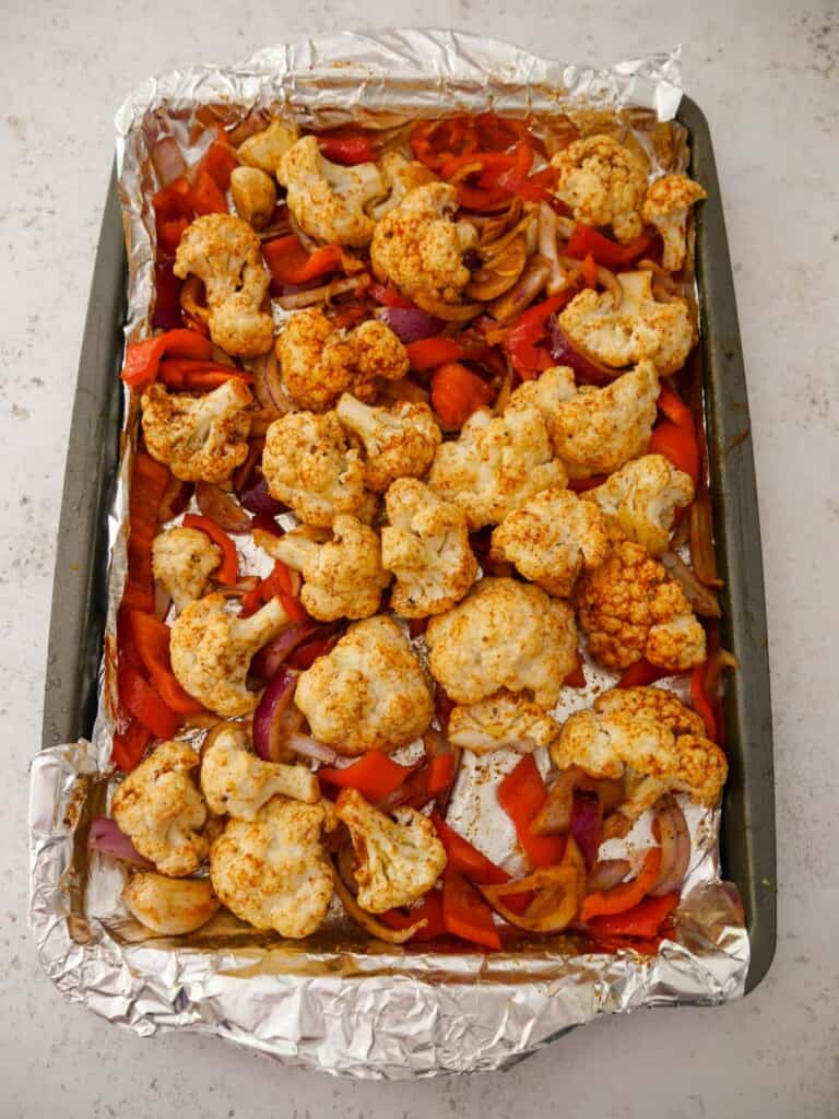 A foil lined baking sheet with cauliflower florets, sliced red onion and red pepper, coated in olive oil and dry spices.