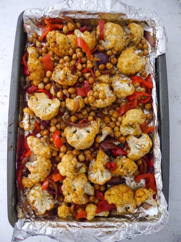 A foil lined baking sheet with part-roasted cauliflower florets, sliced red onion and red pepper with added spiced chickpeas.