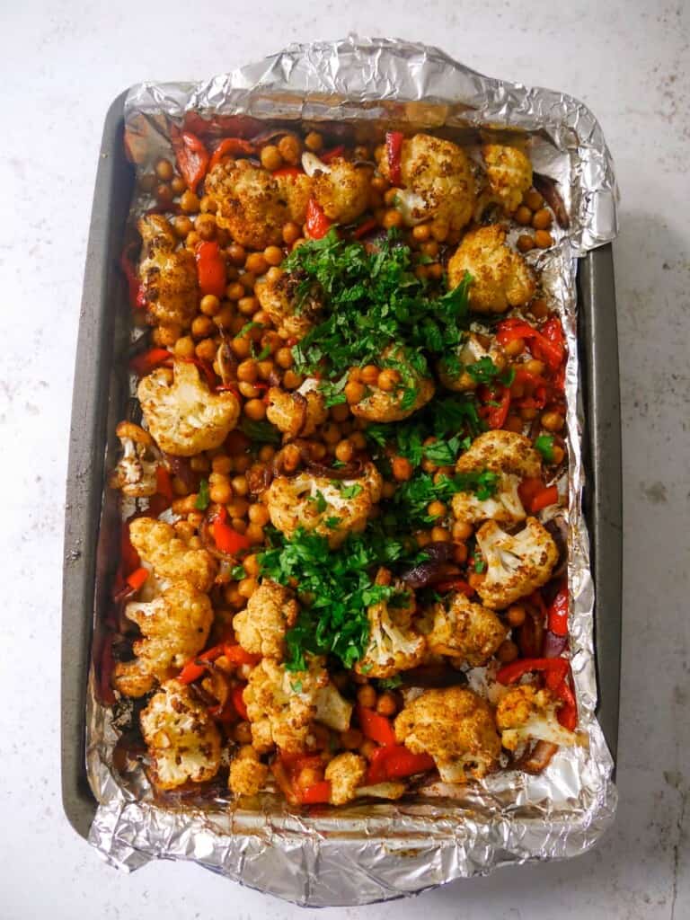 A foil lined baking sheet with roasted cauliflower florets, sliced red onion, red pepper and chickpeas, with added fresh herbs to stir through.