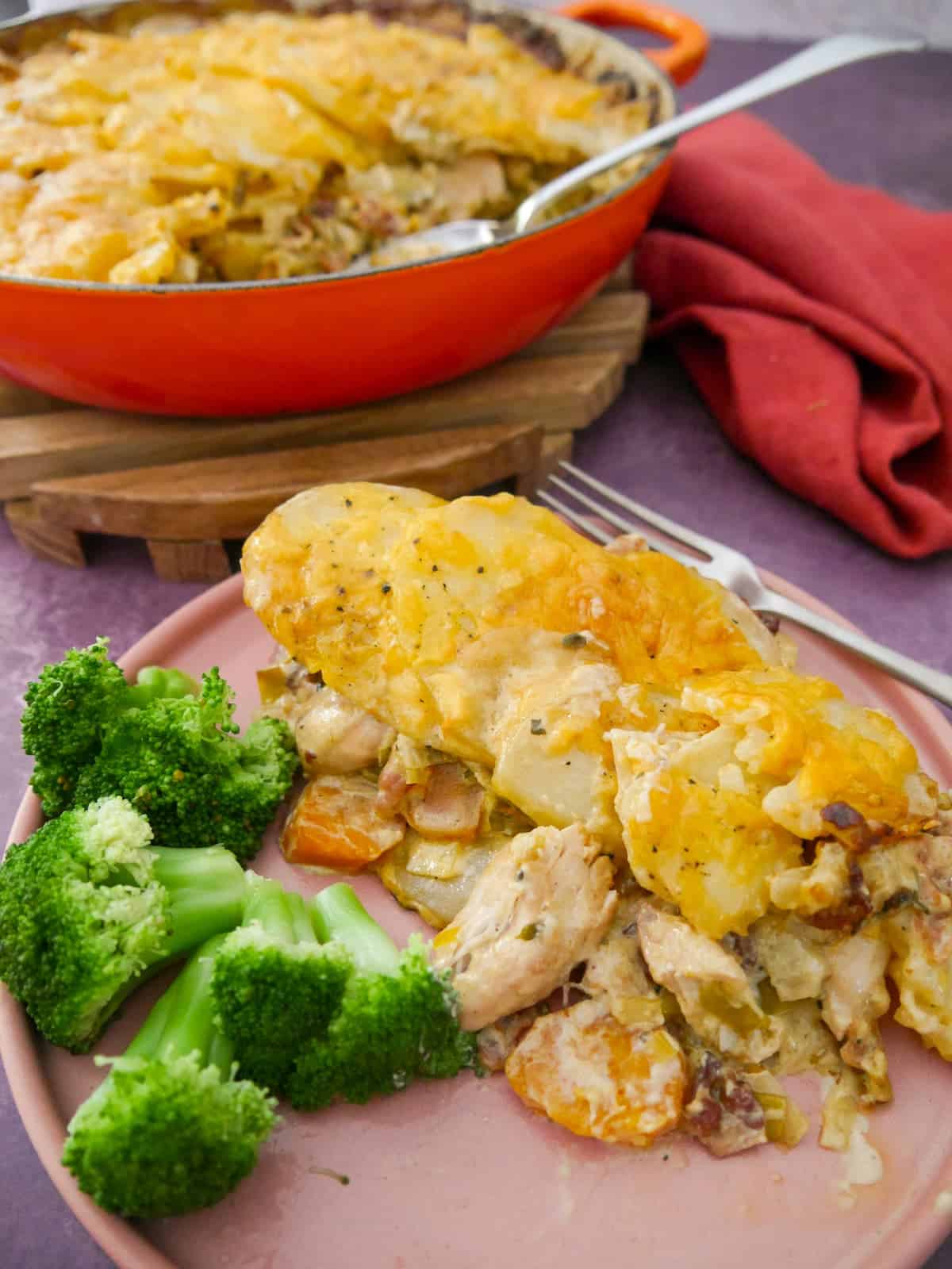 A plate with a serving of chicken and bacon bake in a cider sauce, and steamed broccoli, with a cast iron skillet of the chicken and bacon bake set alongside.