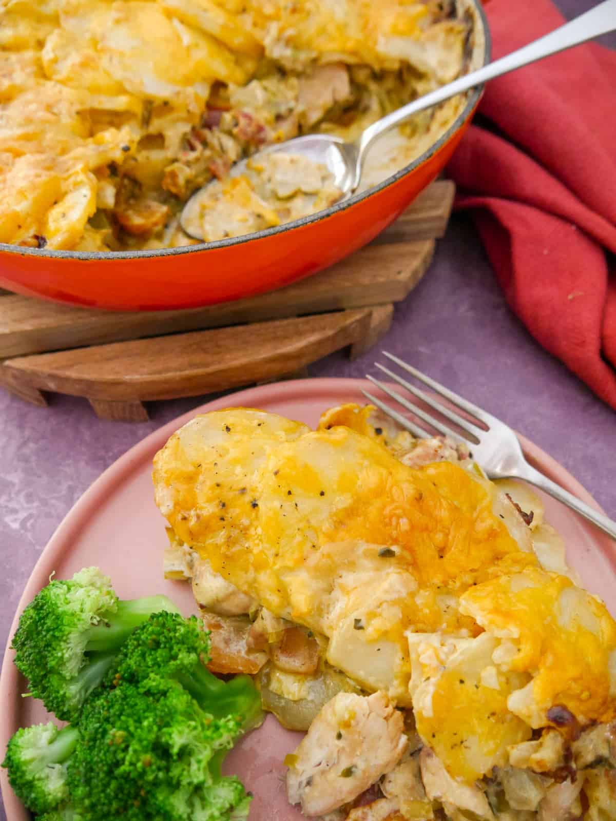 A plate with a serving of chicken and bacon bake in a cider sauce, and steamed broccoli, with a cast iron skillet of the chicken and bacon bake set alongside.