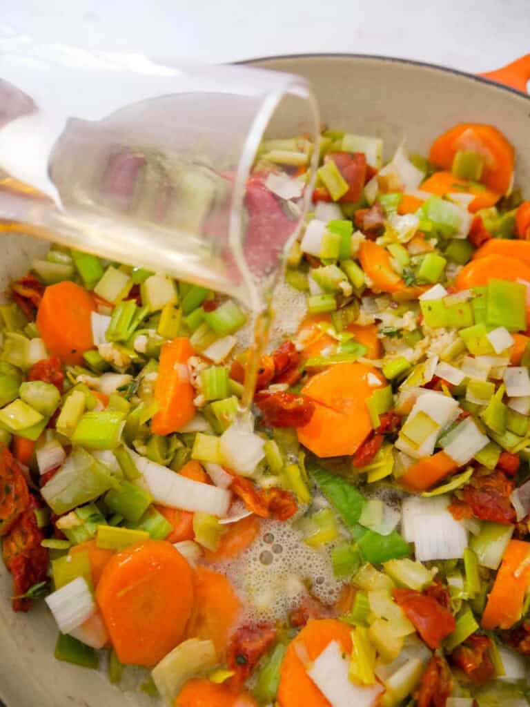 A cast iron skillet of sauteed chicken, bacon and vegetables with cider being poured over.