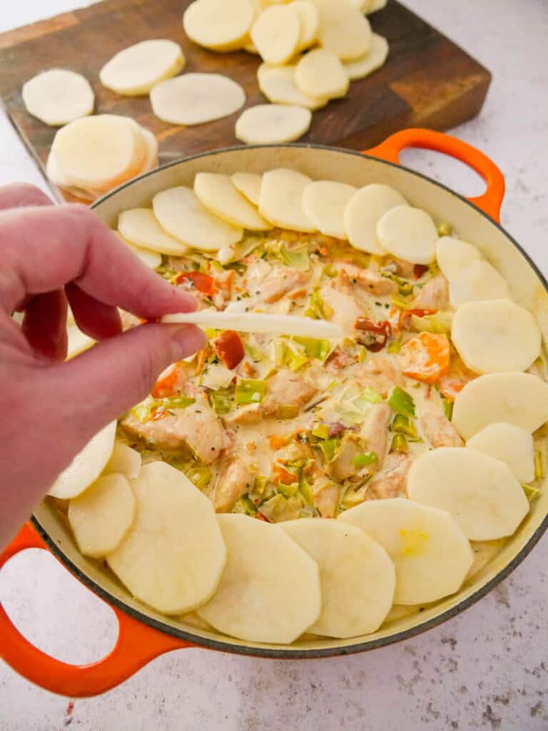 A cast iron skillet filled with chicken and bacon in a creamy cider sauce, with thin slices of potatoes being layered on top.