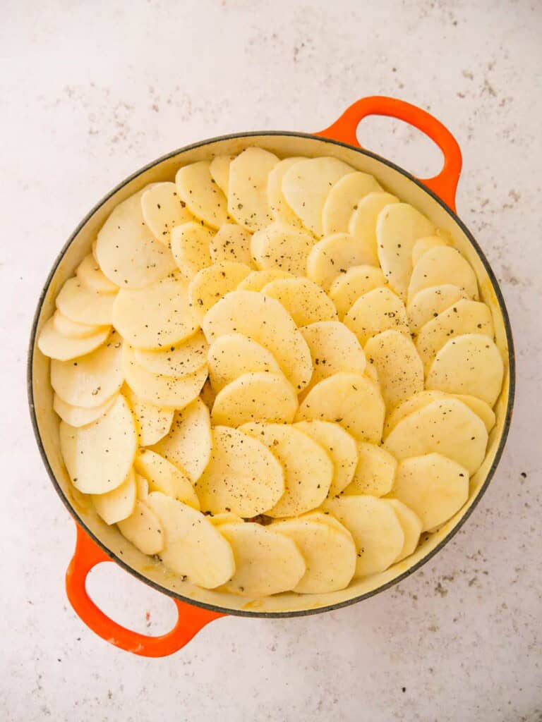 A cast iron skillet filled with chicken and bacon in a creamy cider sauce, topped with a single layer of thinly sliced potatoes seasoned with sea salt and black pepper.