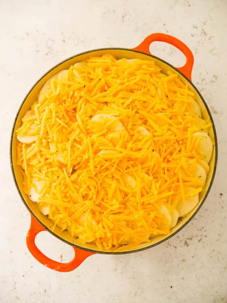 A cast iron skillet, filled with chicken and bacon in a creamy cider sauce, topped with sliced potatoes and grated cheese.