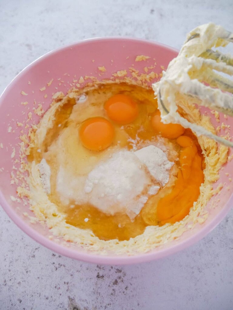 A pink bowl with creamed butter and sugar with added eggs, flour, baking powder, salt and vanilla extract.