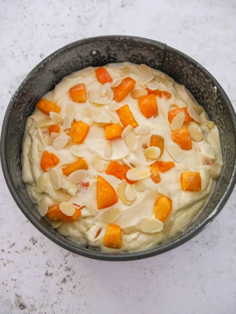 A greased round cake tin, filled with apricot cake mixture and topped with some chopped apricots and flaked almonds.