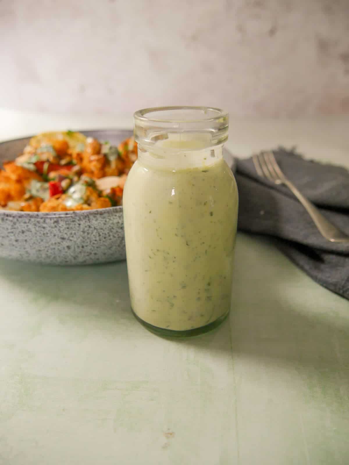 A jar of Greek yogurt lemon herb dressing with a bowl of roasted cauliflower and chickpeas set alongside.