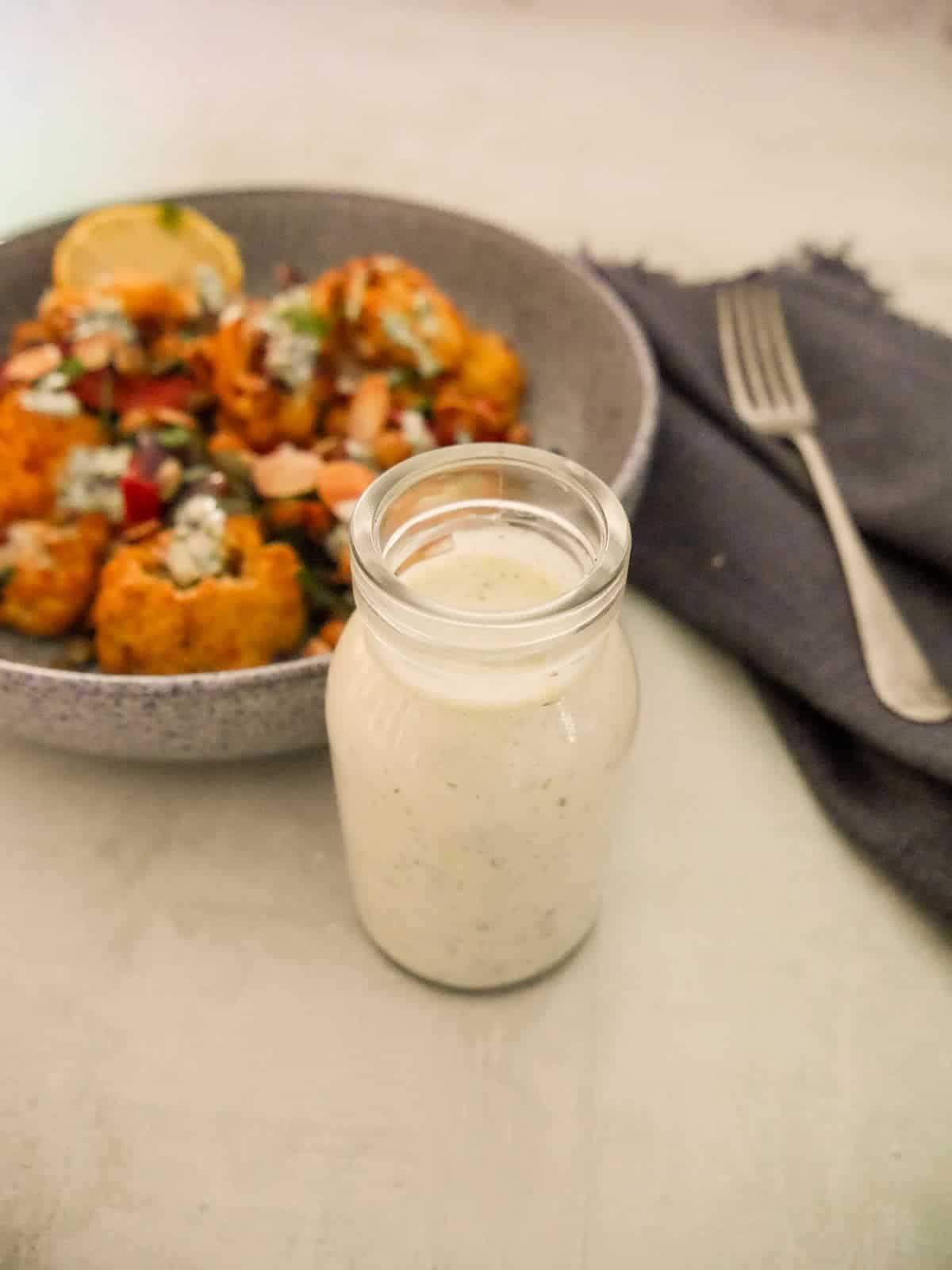 A jar of Greek yogurt lemon herb dressing with a bowl of roasted cauliflower and chickpeas set alongside.