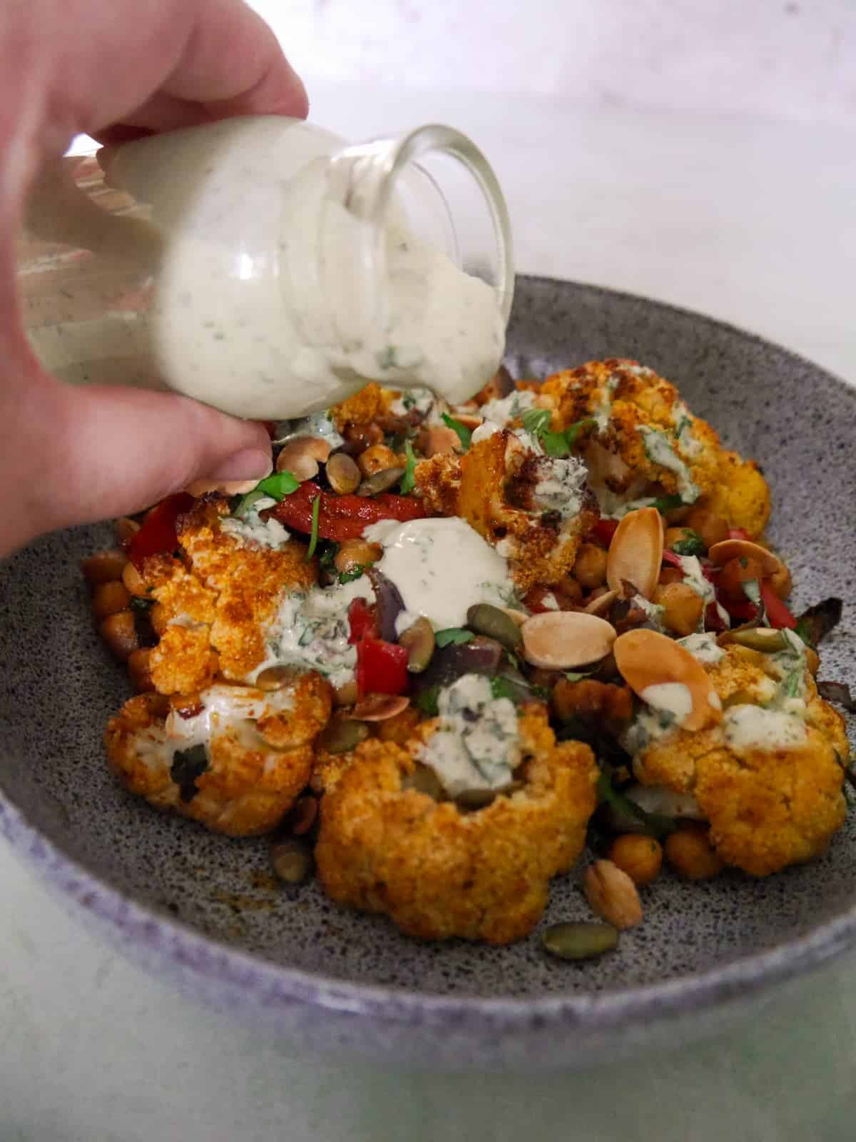 A jar of Greek yogurt lemon herb dressing being poured over a bowl of roasted cauliflower and chickpeas.