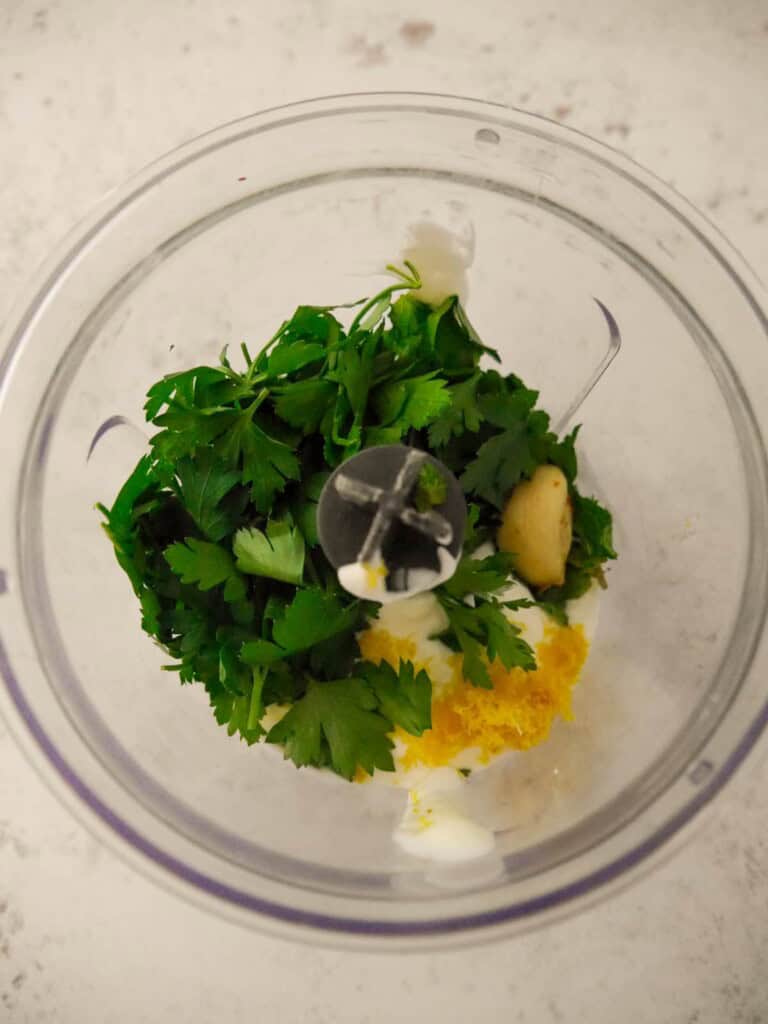 A food processor bowl containing the individual recipe ingredients for making Greek yogurt lemon herb dressing.