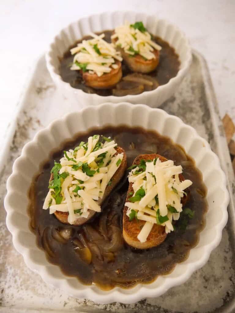 2 bowls of French onion soup, topped with slices of toasted bread and grated gruyere cheese, set on a baking sheet.