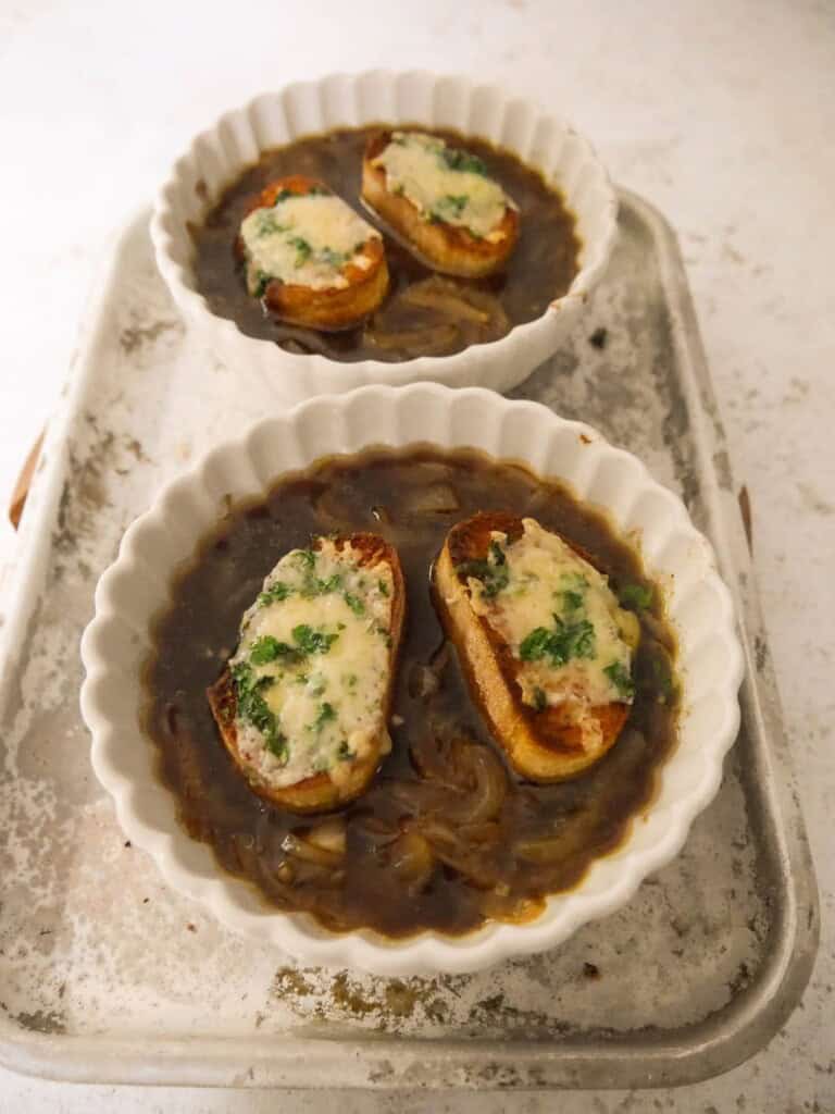 2 bowls of French onion soup, topped with a melted cheese crouton, set on a baking sheet.