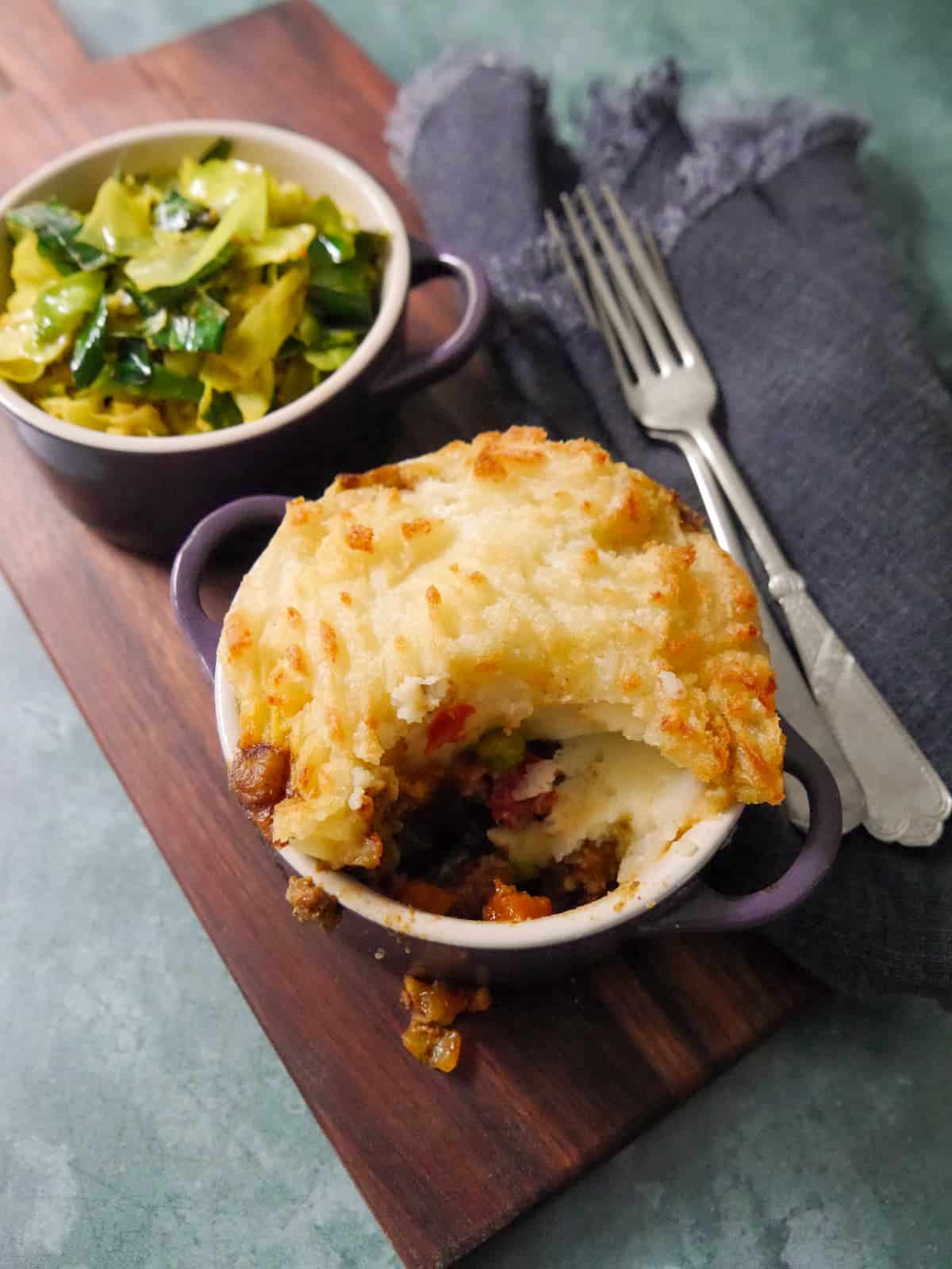 A small dish of masala spiced cottage pie, served with a bowl of mustard spiced cabbage set alongside.