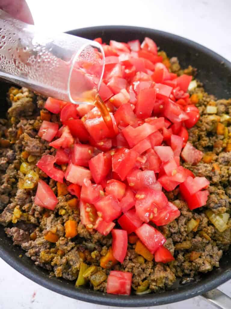 A large frying pan of sauteed and spiced minced beef and vegetables, with added chopped tomatoes and beef stock being poured over.