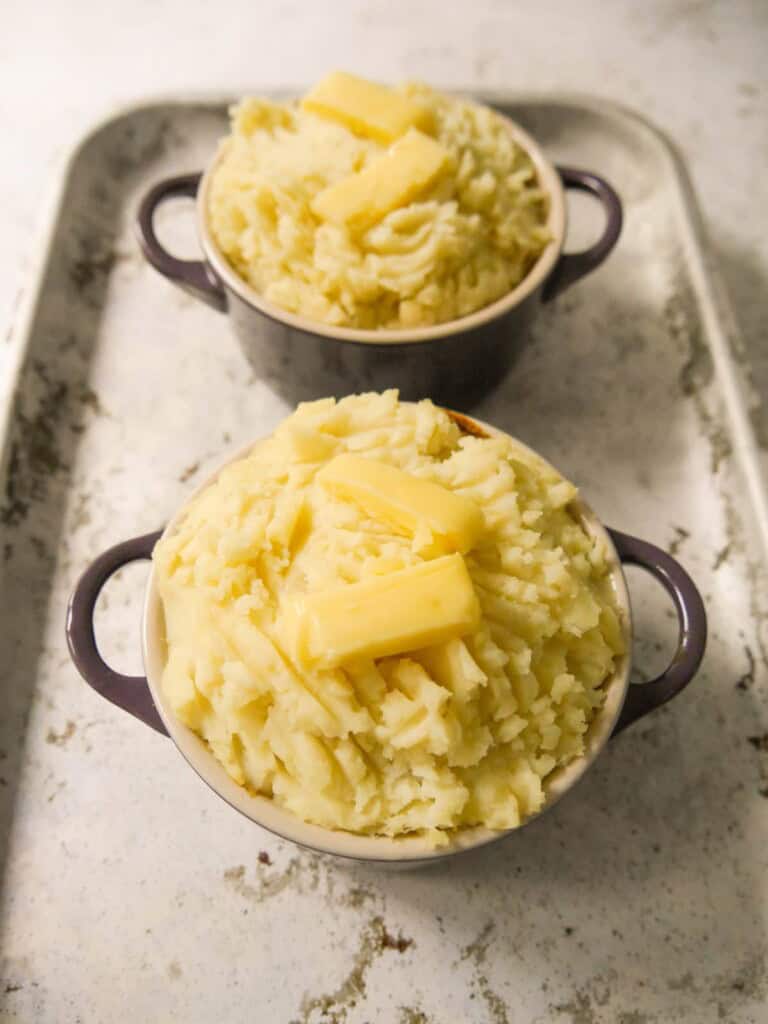 Two dishes of masala spiced cottage pie topped with fluffy mashed potatoes and a knob of butter, ready for the oven.